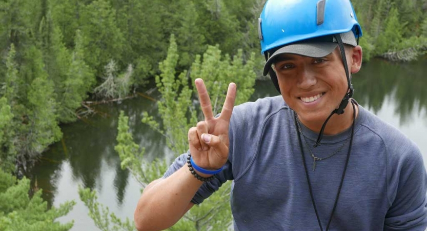 A person wearing a helmet smiles and gives the camera a peace sign. They appear to be at a high elevation. There are green trees and water below them. 